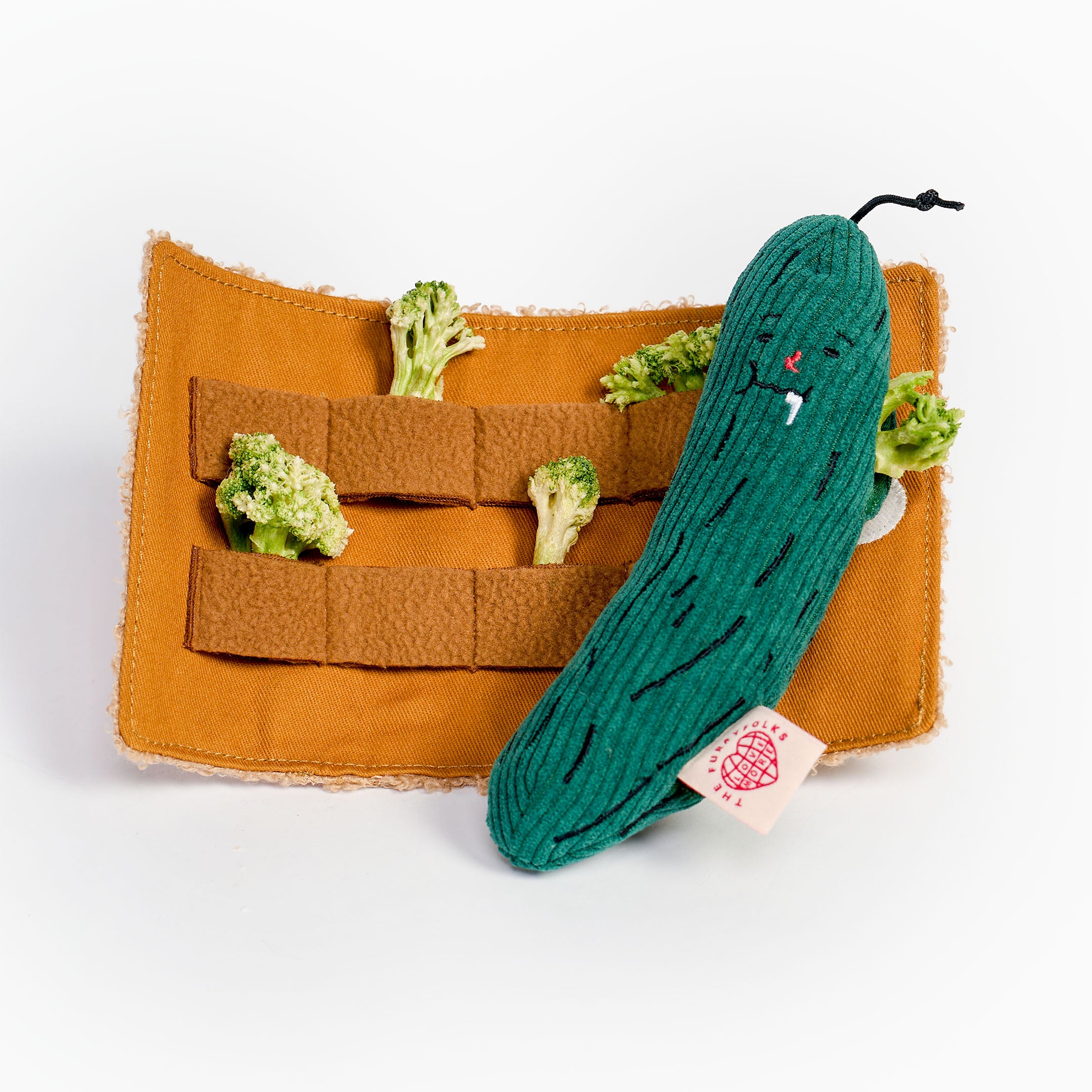 A plush cucumber toy with embroidered facial features and a small tag, resting on an orange fabric organizer insert with small broccoli pieces inside, photographed on a clean white background.