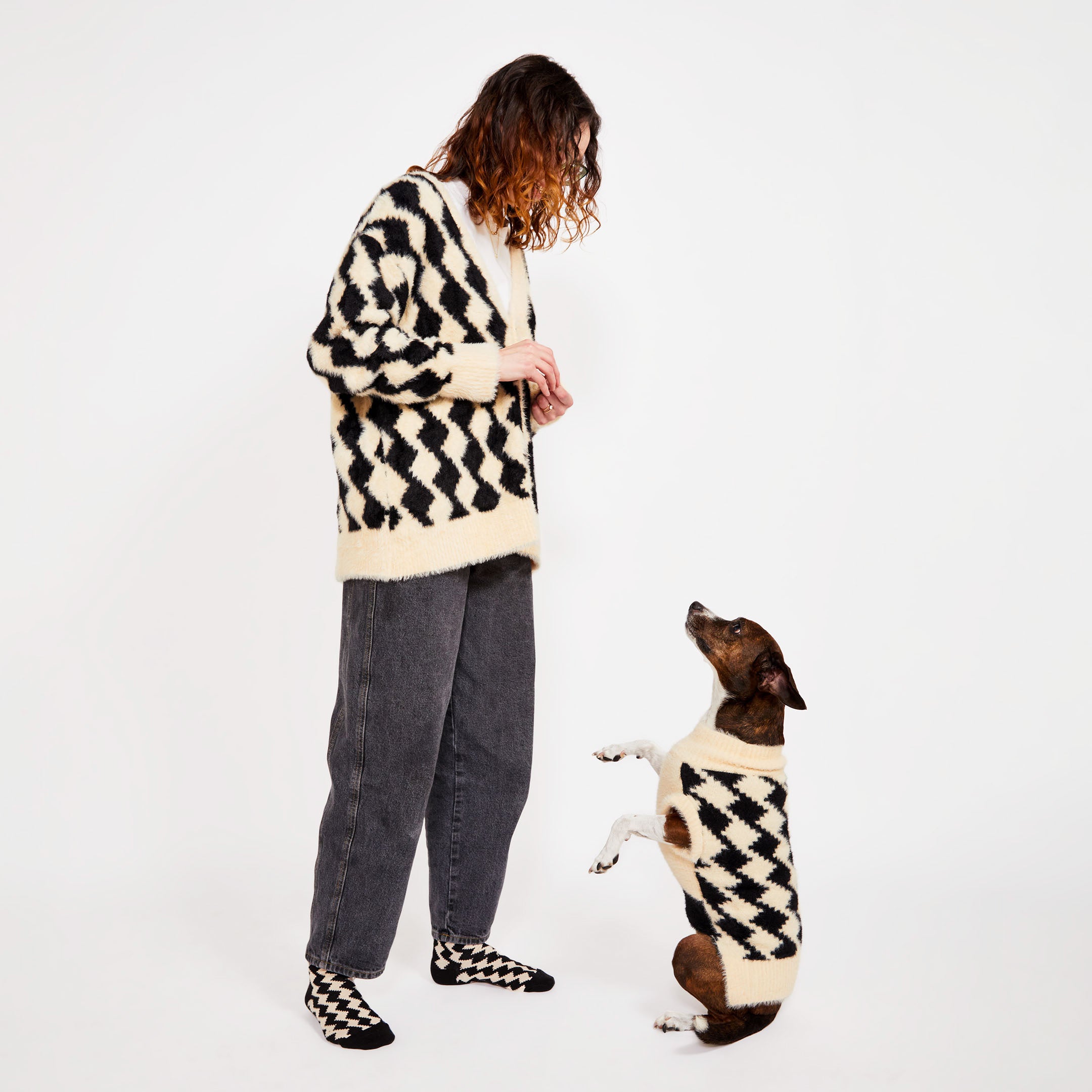 A woman standing with a brown dog who is standing on its hind legs. Both are wearing matching black and cream-colored knitwear with a diamond pattern. The woman is also wearing matching socks with black and cream diamonds.