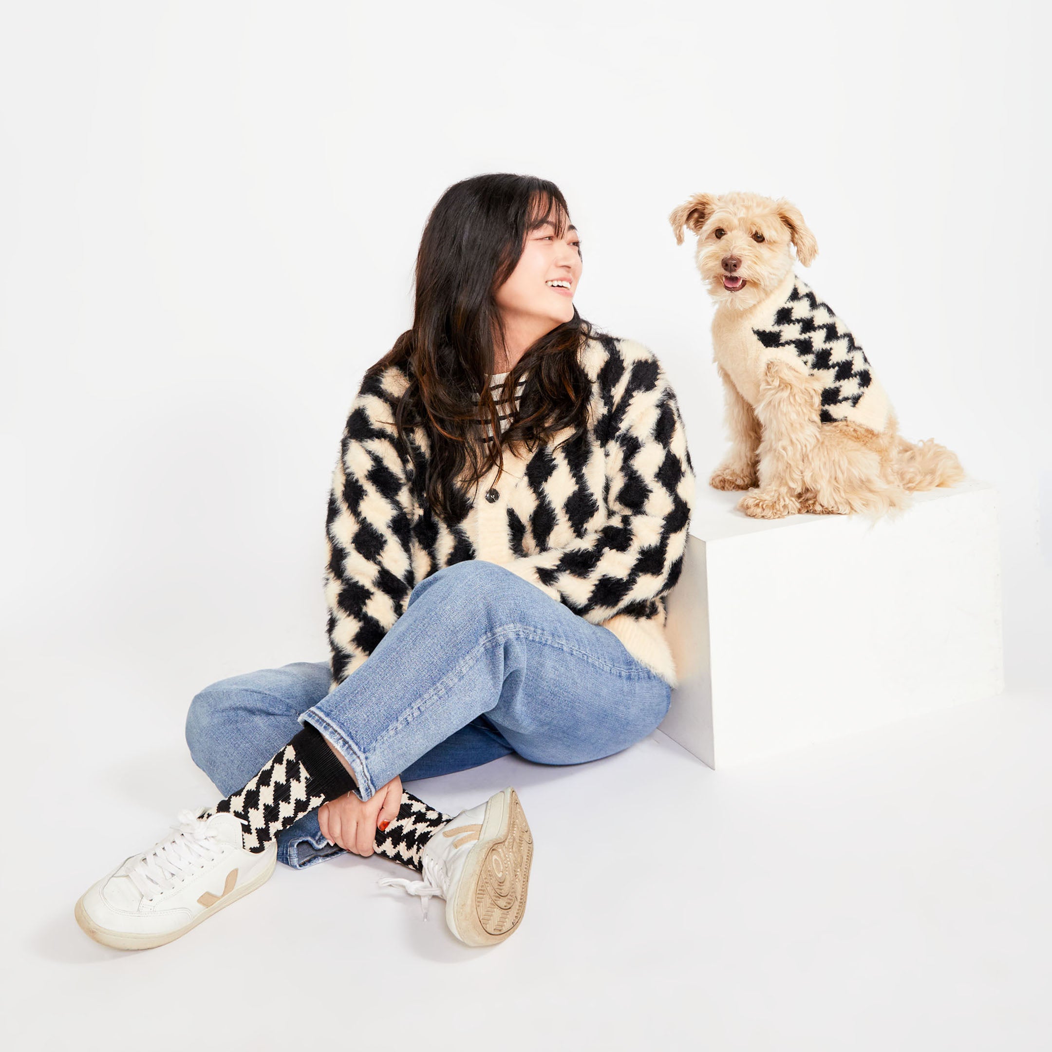  A woman sitting on the floor with her dog beside her. Both are wearing matching black and cream-colored sweaters with a diamond pattern. The woman is also wearing matching socks, completing the cozy look.