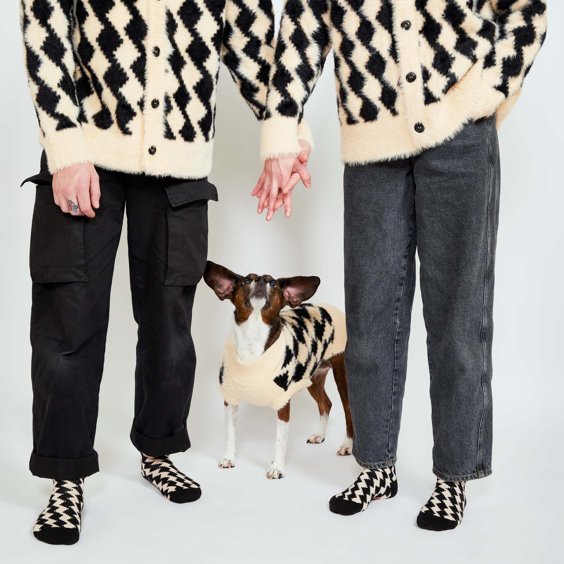 Two people standing, holding hands, while a brown dog looks up at them. All three are dressed in matching black and cream-colored knitwear with a diamond pattern.