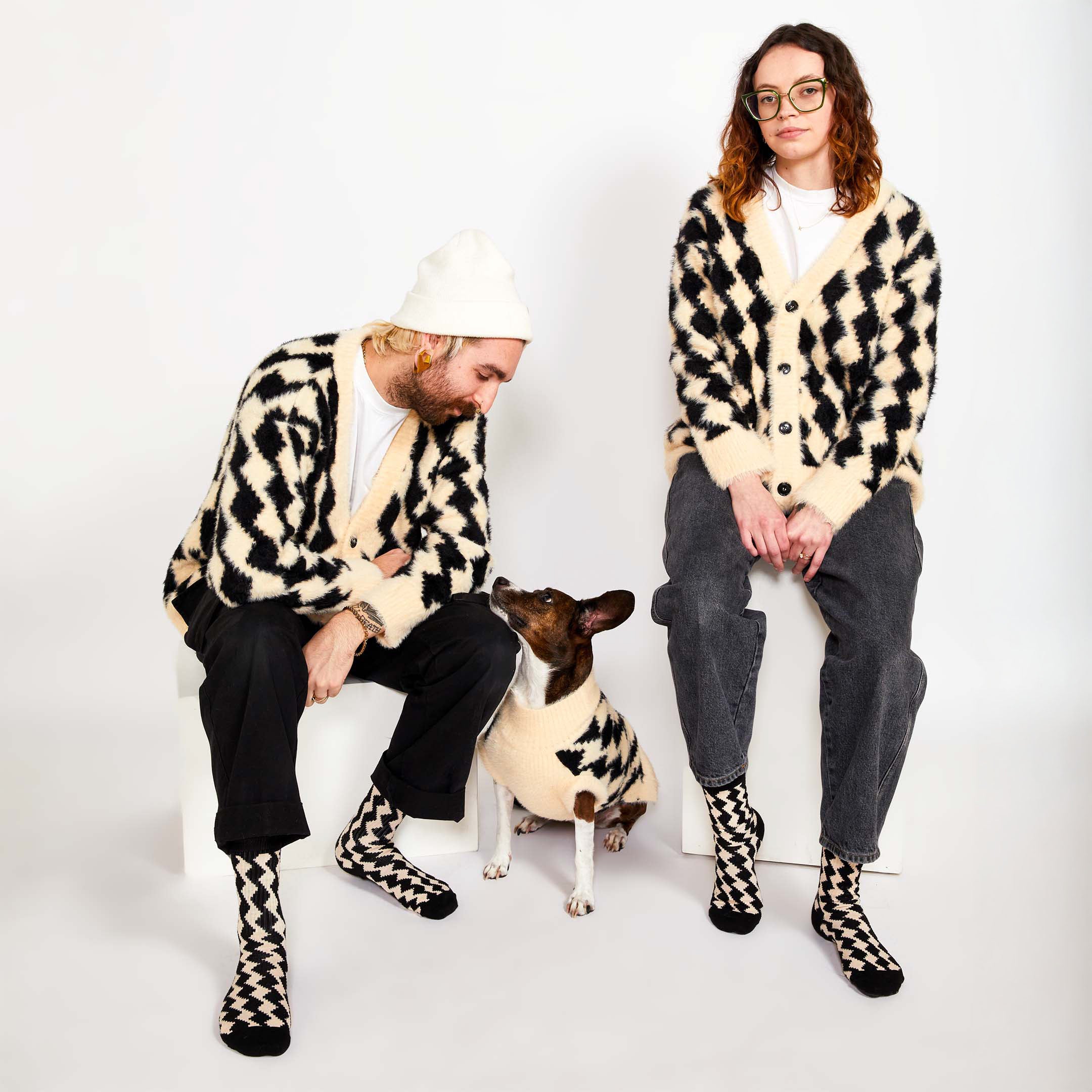  Two people seated with a brown dog between them. All three are wearing matching black and cream-colored sweaters with a diamond pattern. The people are also wearing matching socks.