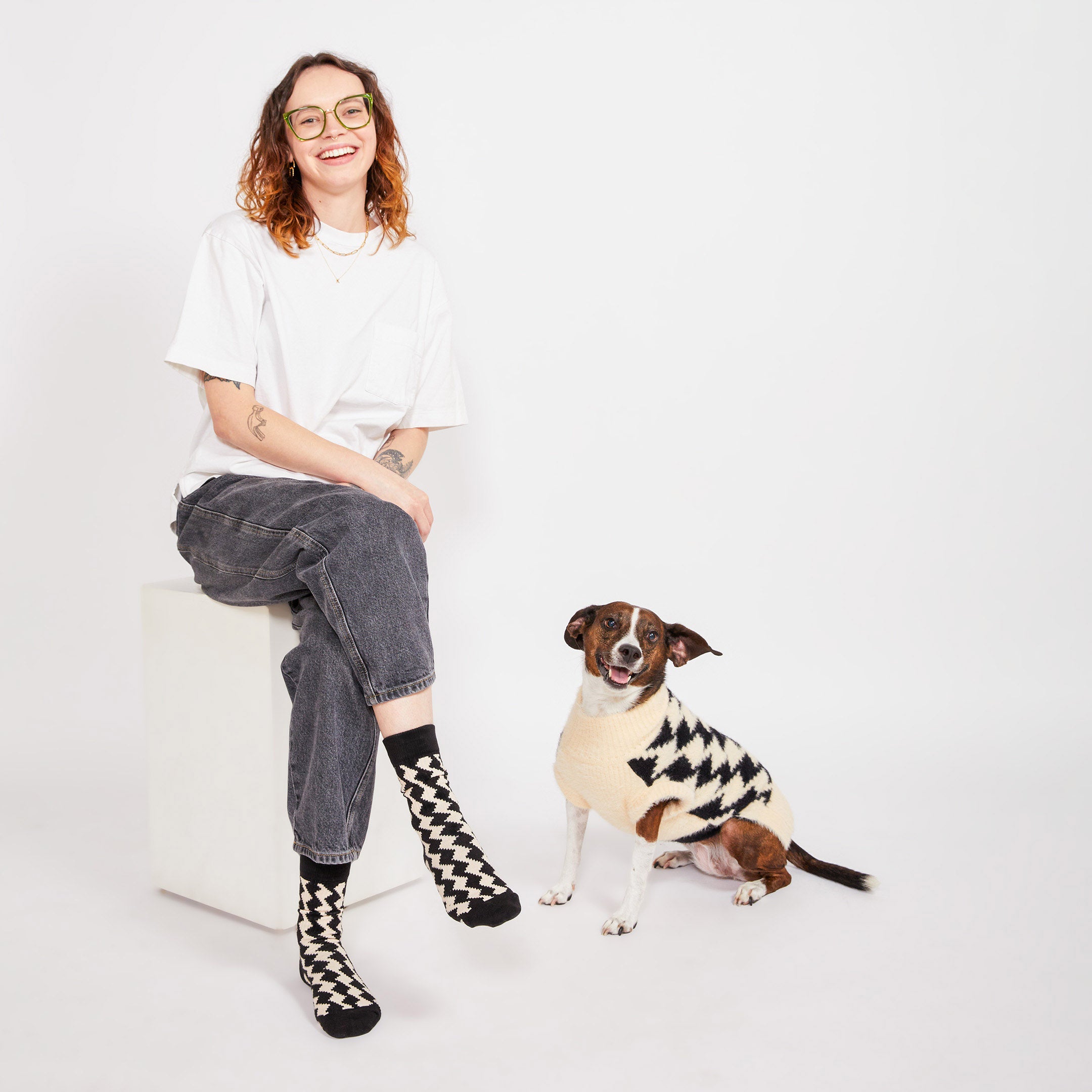 A woman sitting on a white block, smiling at the camera, wearing black and cream socks with a diamond pattern, while her brown dog, dressed in a matching sweater, sits beside her.