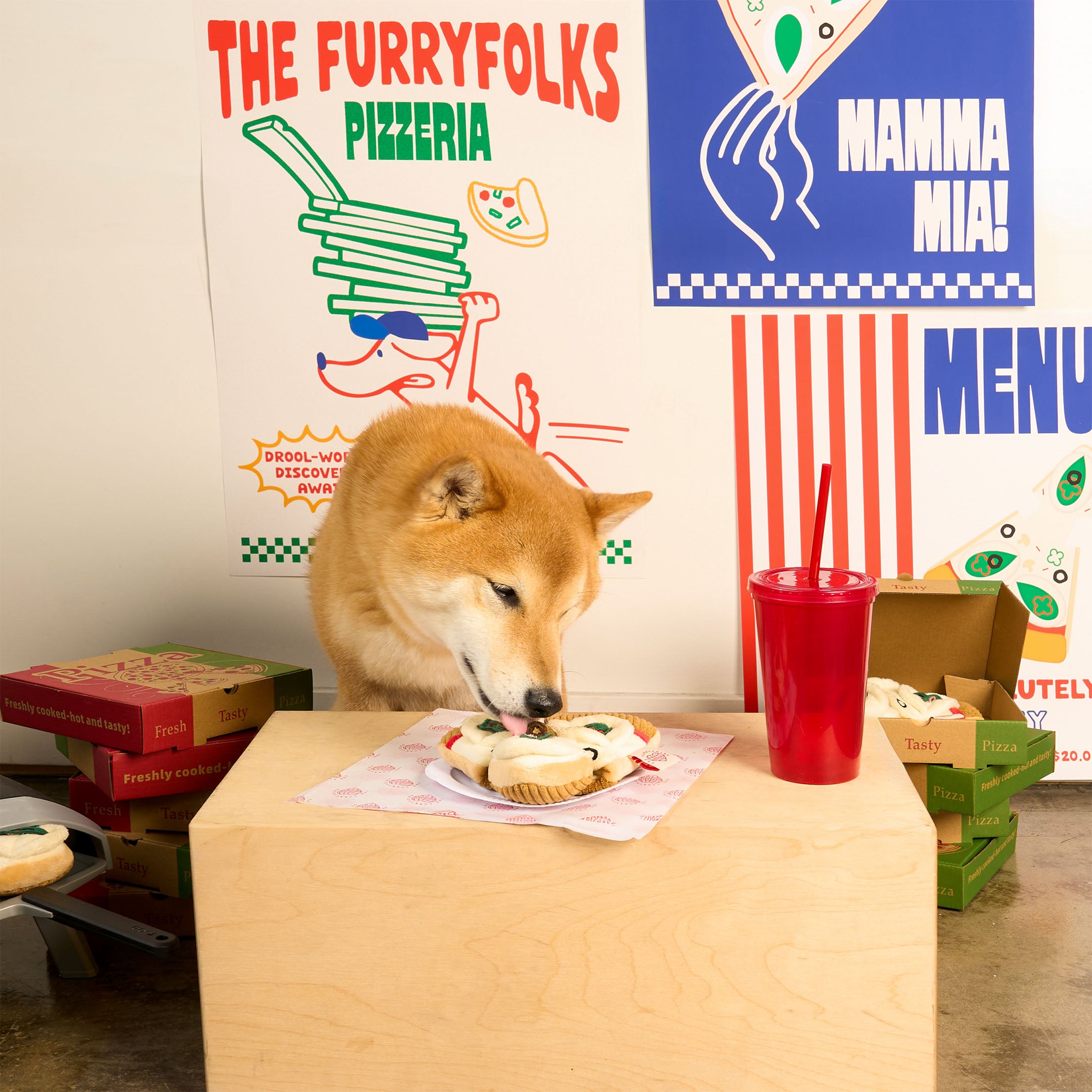 Shiba Inu sniffing a plush pizza slice nosework toy on a wooden table, surrounded by playful pizzeria-themed decor. A fun and whimsical setup featuring the furryfolks’ interactive dog toy.