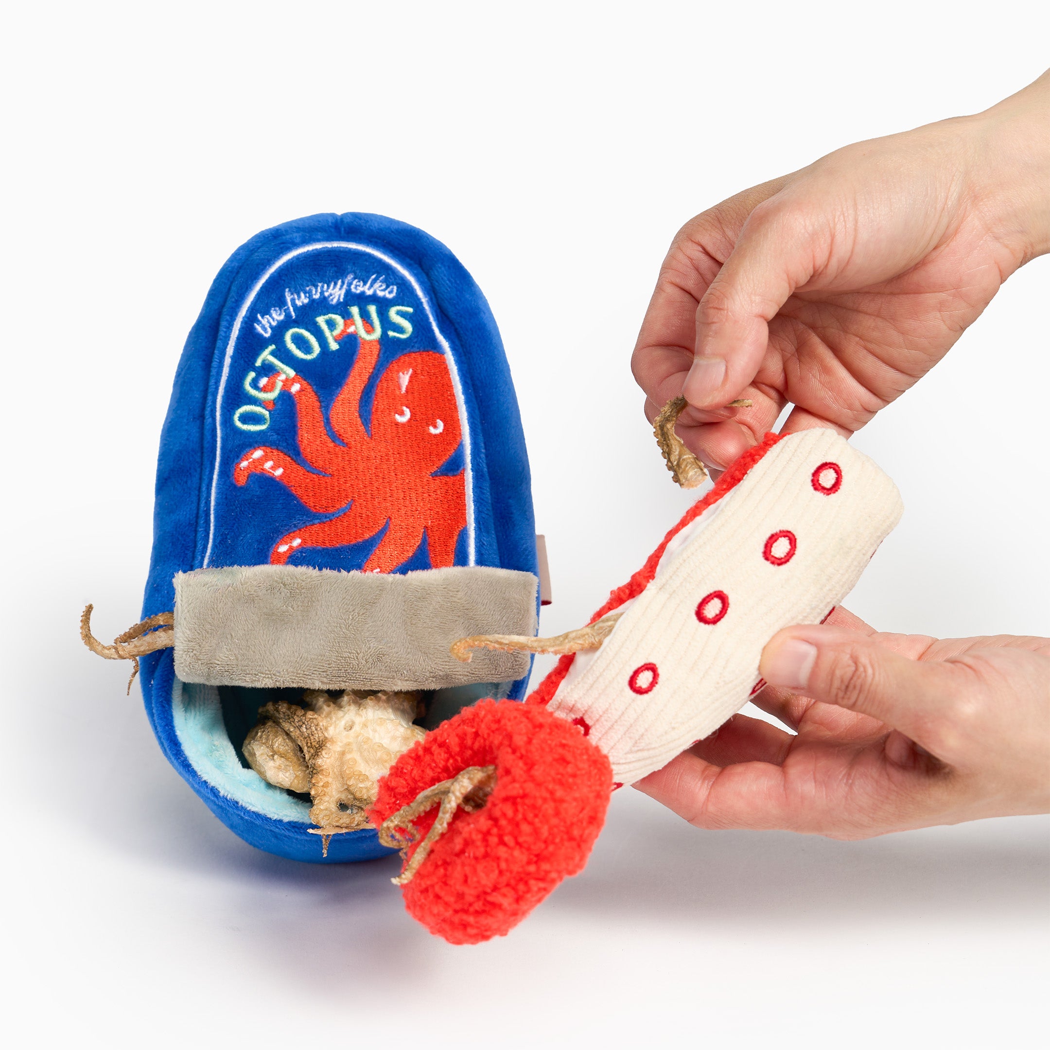 A blue plush octopus-shaped pet toy with an open flap reveals beige stuffing and a red pom-pom tail. A person's hands are holding a matching beige and red plush octopus tail toy against a white background.