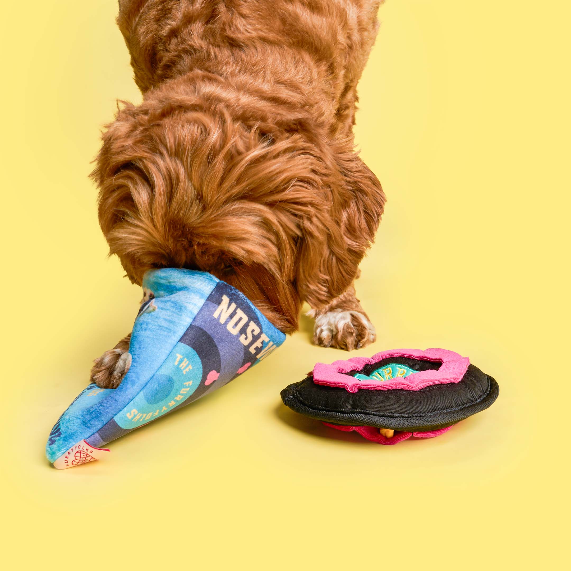 Brown dog buries its nose deep inside the record sleeve part of the “Spin ‘N’ Sniff” toy, with the vinyl-shaped nosework disc lying nearby on a yellow background.