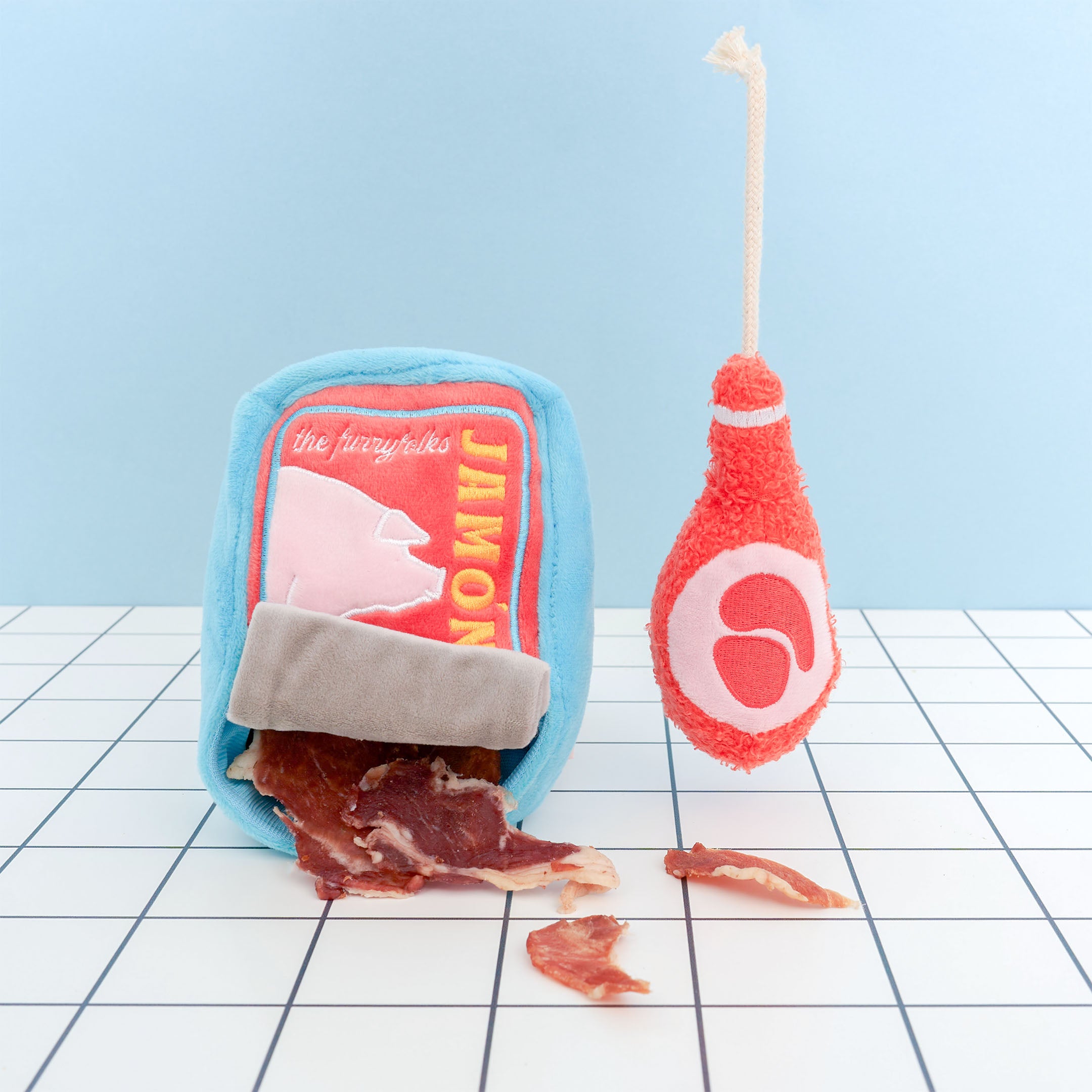 A plush dog toy shaped like a can of jamón (ham) sits on a grid-patterned surface. The blue toy has a red front label with a pink pig silhouette and the word “JAMÓN” in yellow. A gray flap at the bottom resembles a can tab, with fabric pieces resembling ham spilling out. Next to the can is a plush ham leg toy in red with a white rope attached at the top, mimicking a ham hock.