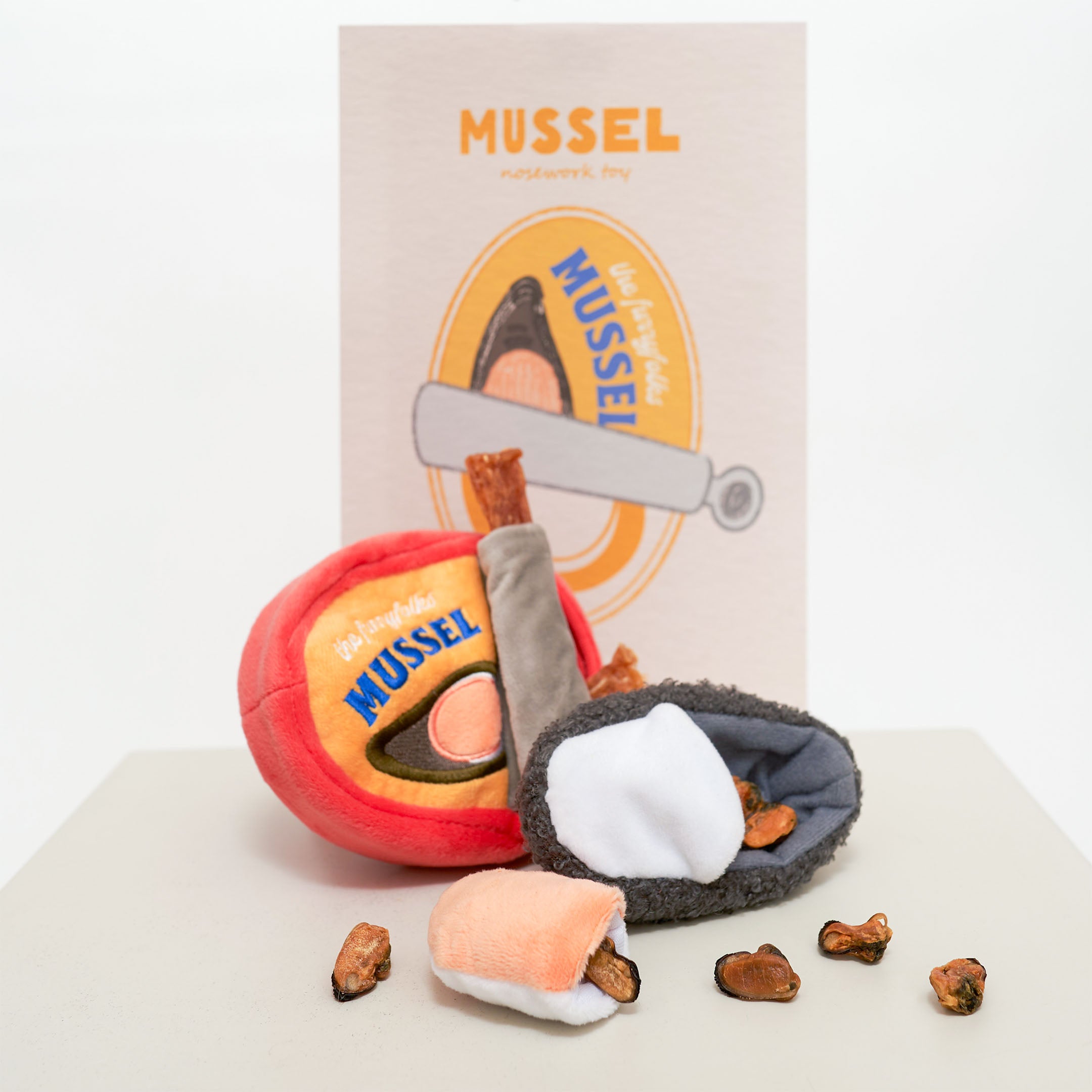 The image shows a dog nosework toy set called “Mussel.” The set includes a red plush container with a yellow label, a grey mussel shell toy, and small brown bone-shaped treats. In the background, there is a card with “Mussel” branding.