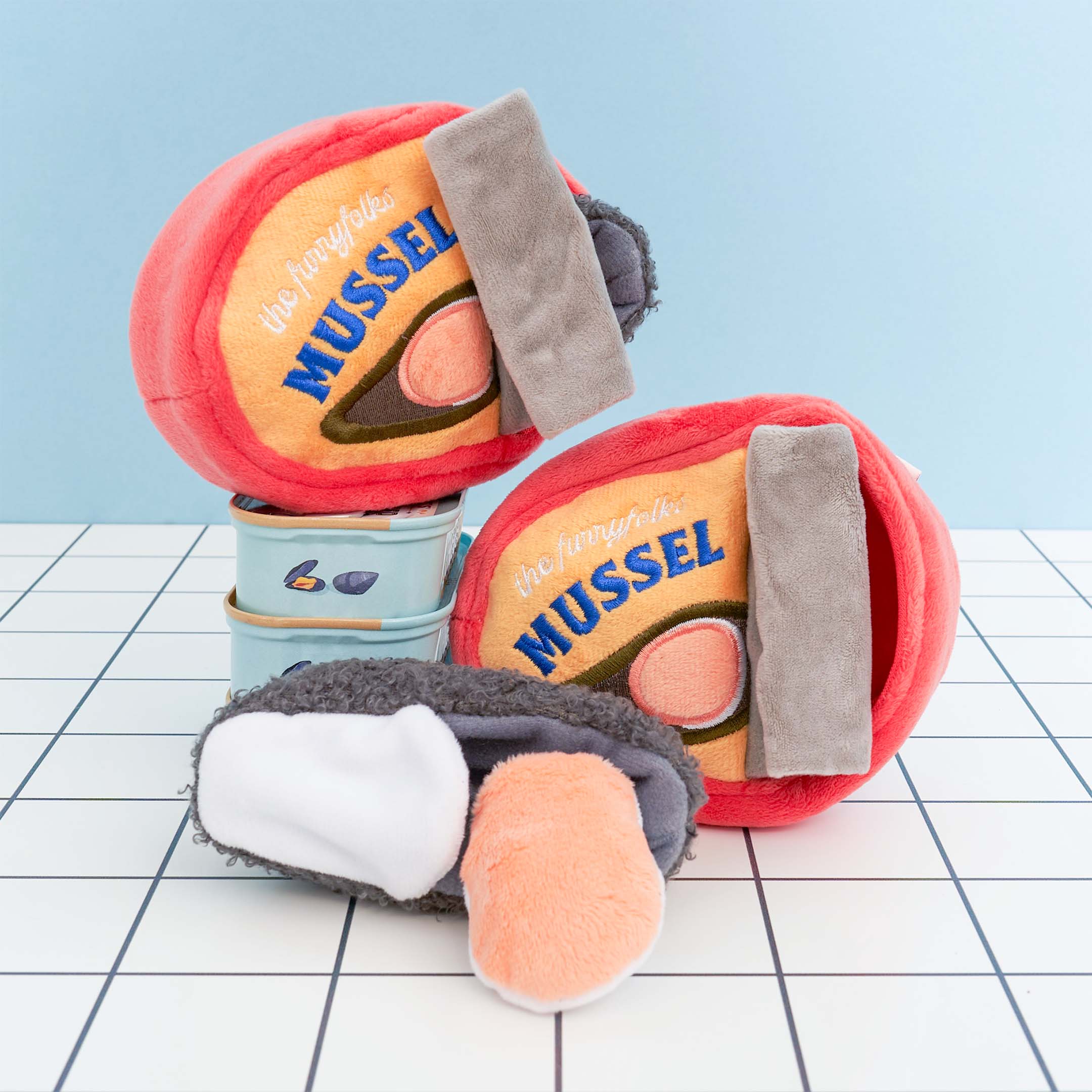 The image features two plush dog toys shaped like cans of mussels and one plush mussel toy. The can-shaped toys are red with yellow labels featuring an illustration of a mussel and the word “MUSSEL” in blue letters. Each toy has a gray flap resembling a pull tab. One of the plush mussels, which is gray with a white and orange interior, is placed in front of the cans. The toys are designed for dog nosework activities and are set against a grid-patterned surface with a light blue background.