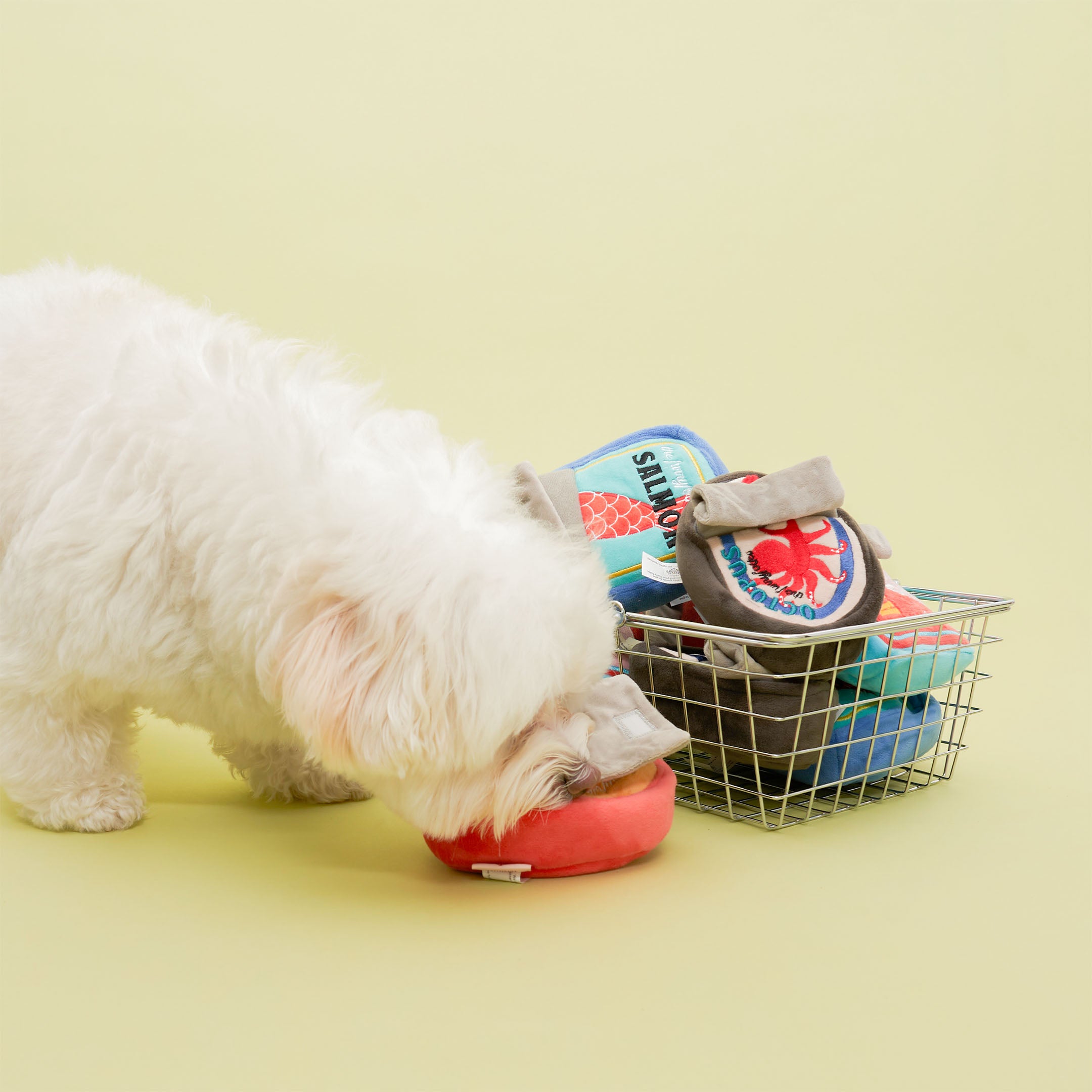 A small, fluffy white dog is sniffing a red plush toy on the ground. Next to the dog is a wire basket filled with various plush toys designed to look like cans of different foods. The toys have colorful labels, including one that looks like a can of salmon and another that resembles a can with a crab label. The scene is set against a light yellow background.