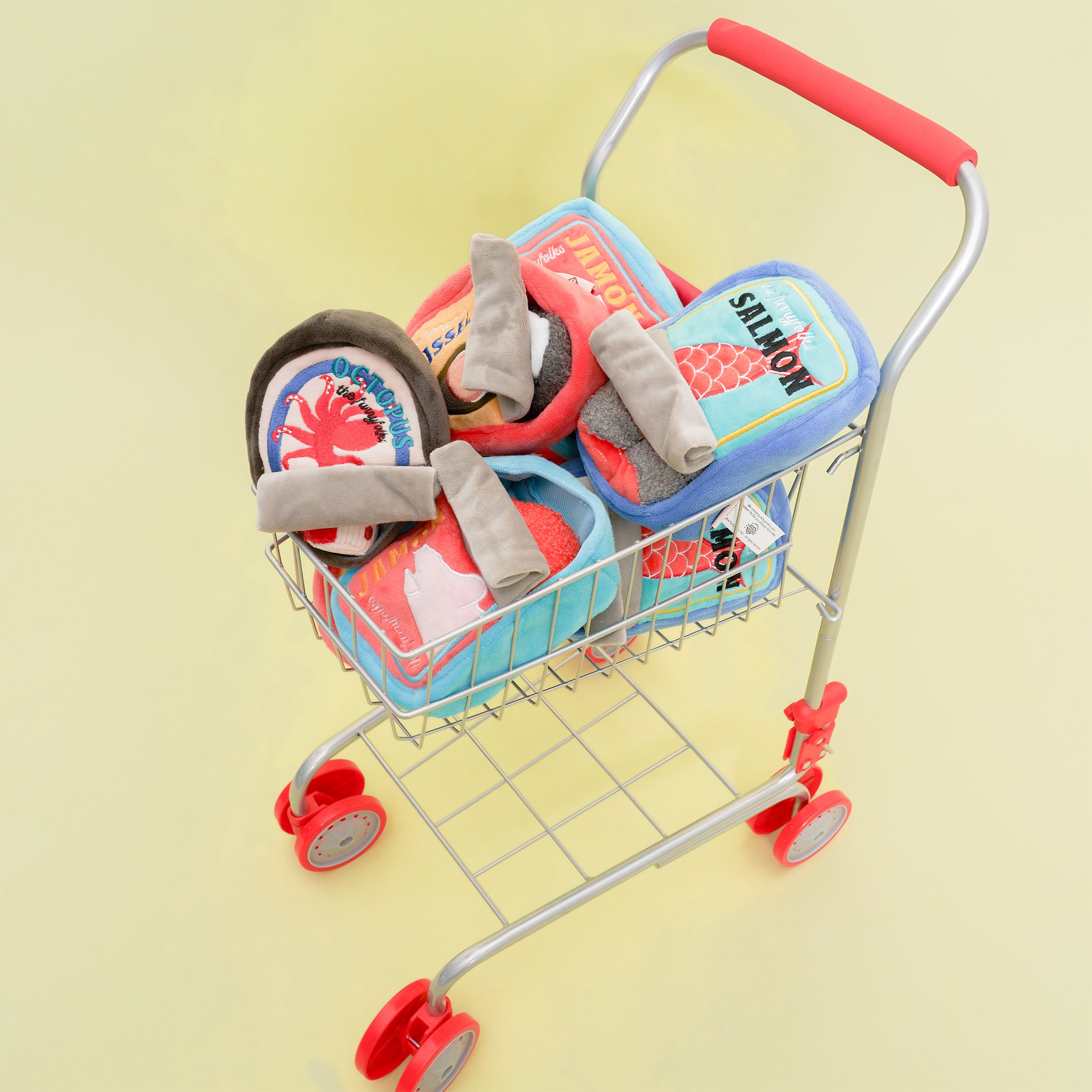 A small wire shopping cart with a red handle is filled with various plush dog toys designed to look like canned foods. The toys have colorful labels and are shaped like cans, featuring designs such as a red crab labeled “Octopus,” a red pig labeled “JAMÓN,” and a fish labeled “SALMON.” The shopping cart is set against a light yellow background.