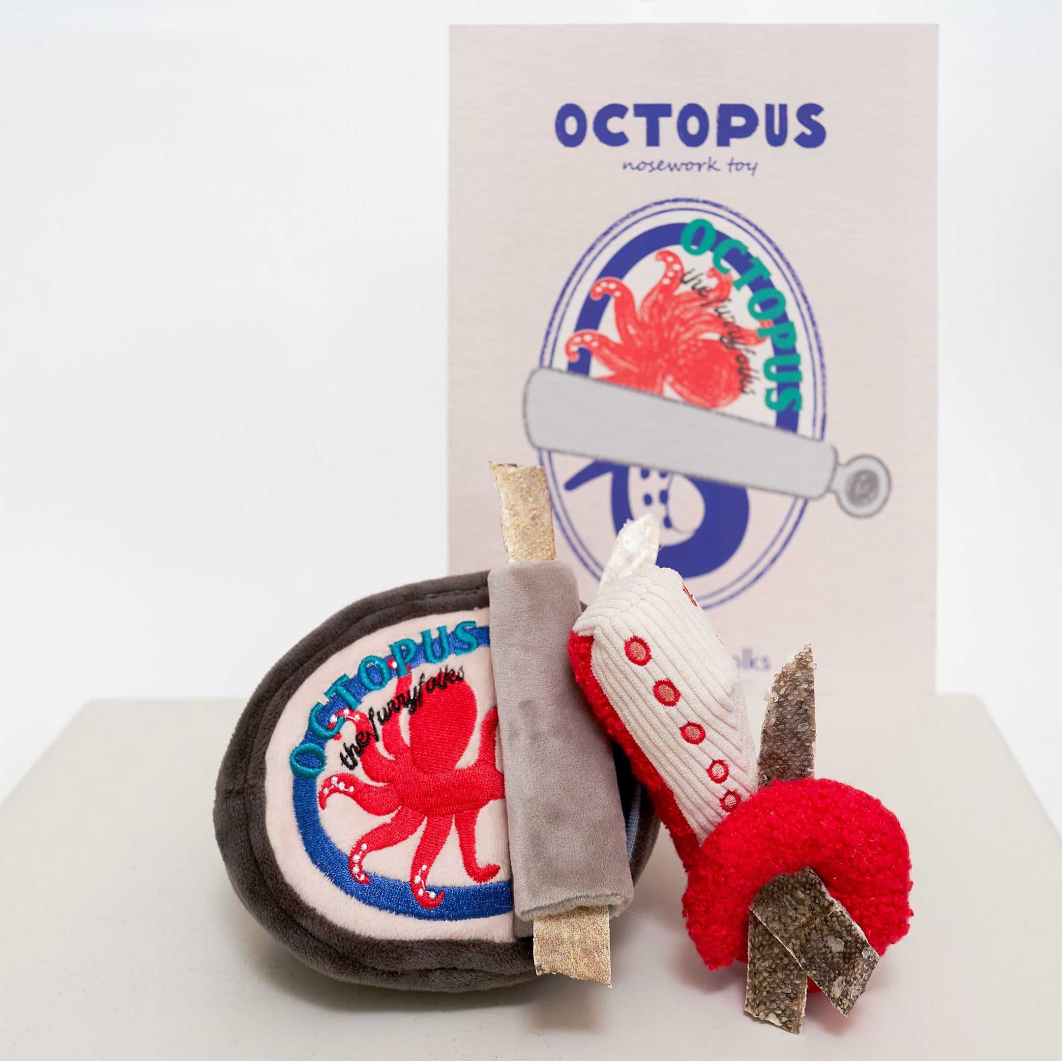 The image shows a dog nosework toy set called “Octopus.” The set includes a grey plush container with a red octopus design, a grey roller toy, and a red and white octopus leg toy. In the background, a card with “Octopus” branding is visible.