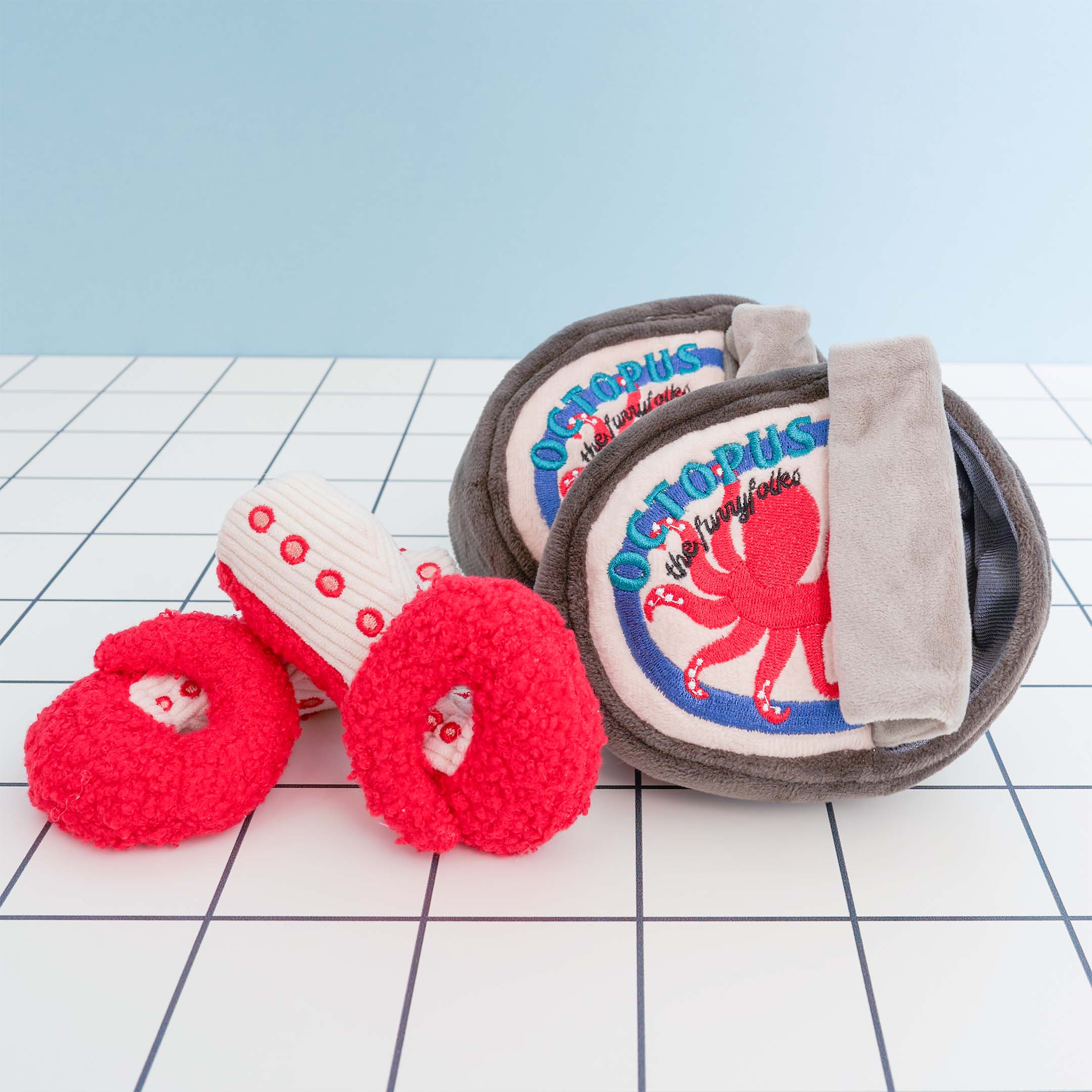 The image features two plush dog toys shaped like cans of octopus and one plush octopus tentacle toy. The can-shaped toys are dark brown with cream-colored labels featuring a red octopus illustration and the word “OCTOPUS” in blue letters. Each can toy has a gray flap resembling a pull tab. In front of the cans are two plush octopus tentacles, one red and one white with red spots. The toys are designed for dog nosework activities and are set against a grid-patterned surface with a light blue background.