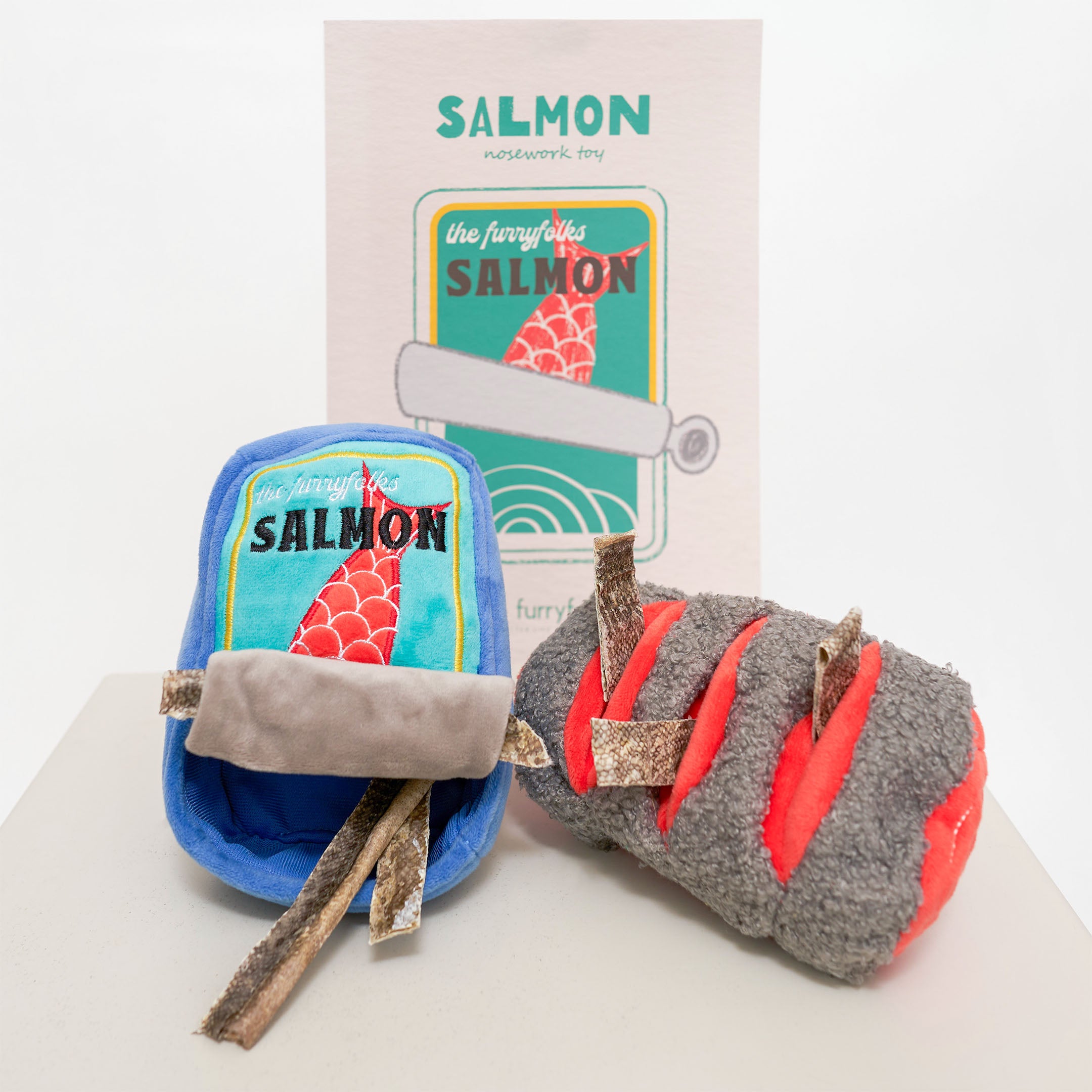 The image shows a dog nosework toy set called “Salmon.” The set includes a blue plush container with a label featuring a red fish and the words “Salmon” and “the furryfolks.” Also included is a grey plush salmon fillet toy with red sections. In the background, a card with similar “Salmon” branding is visible.