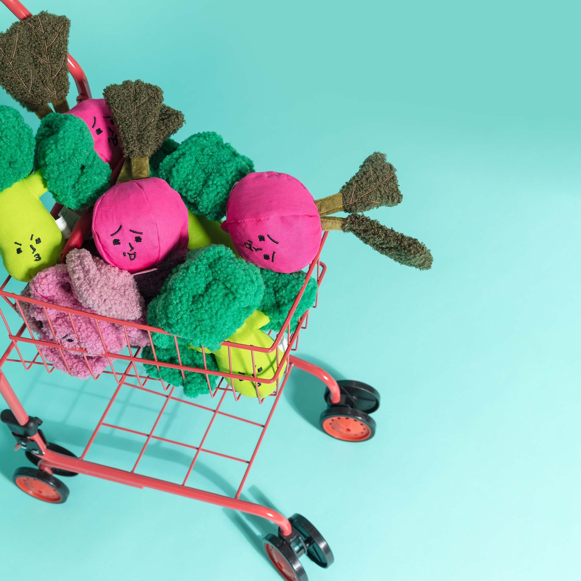 The image shows a miniature shopping cart overflowing with colorful plush dog toys designed as vegetables with expressive faces. There are broccoli, radish, and other garden variety shapes, each with a whimsical character. The scene is set against a bright turquoise background, which enhances the playful and vibrant appearance of the toys, suggesting a fun and engaging playtime for dogs.