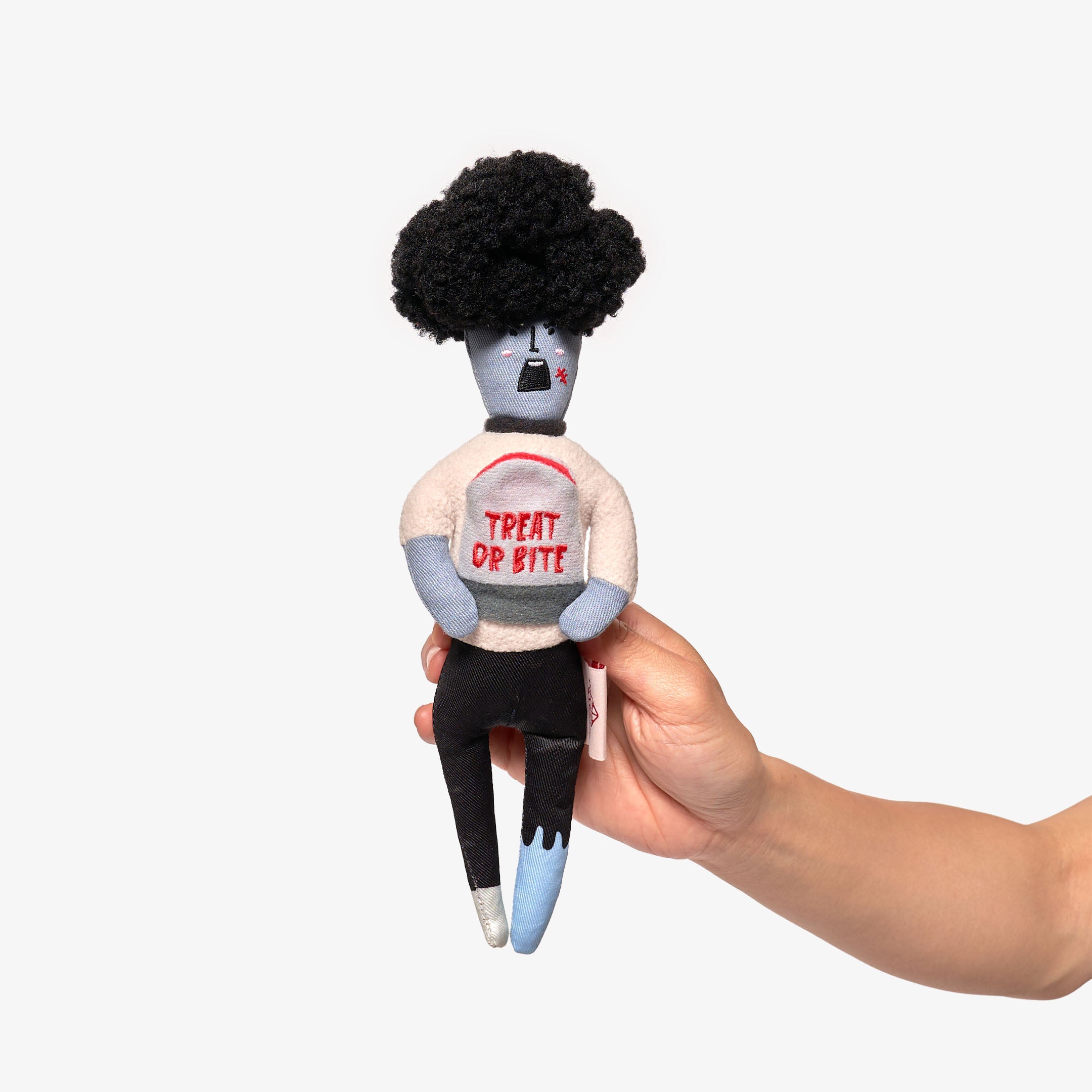 The image displays a hand holding a plush dog toy designed to resemble a zombie. The toy has a gray and blue body with a black, afro-style hairpiece and features the playful phrase "TREAT OR BITE" on its chest. This dog toy is intended for nosework, where treats can be hidden inside for pets to find, providing both mental stimulation and entertainment. The light background contrasts with the toy, highlighting its details and whimsical design.