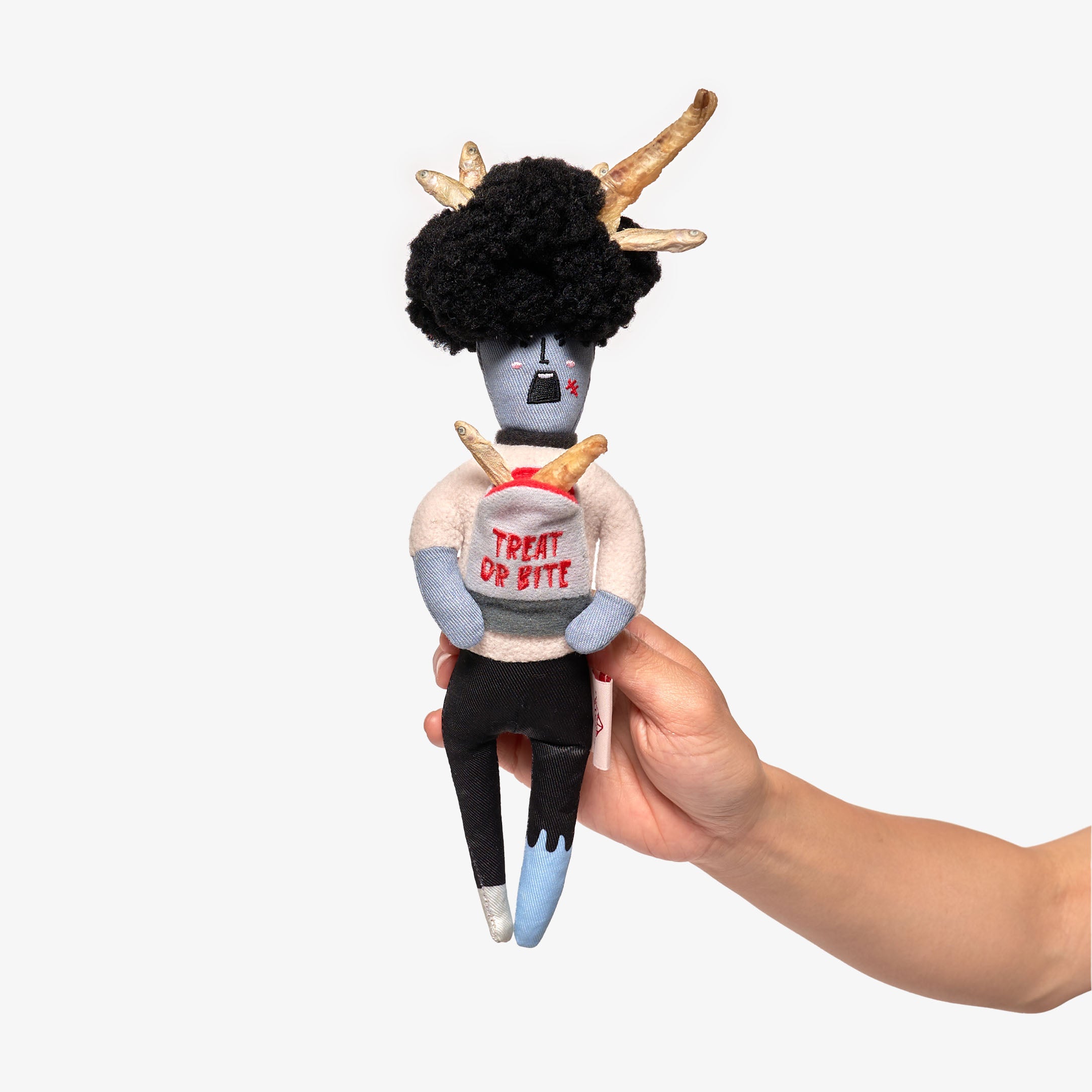 The photo displays a hand holding a whimsical dog toy with a zombie theme, featuring antlers, a black curly hairdo, and a slogan "TREAT OR BITE" on its torso. This interactive toy is designed for nosework, encouraging dogs to sniff out hidden treats. The light background highlights the toy's playful and creative design.