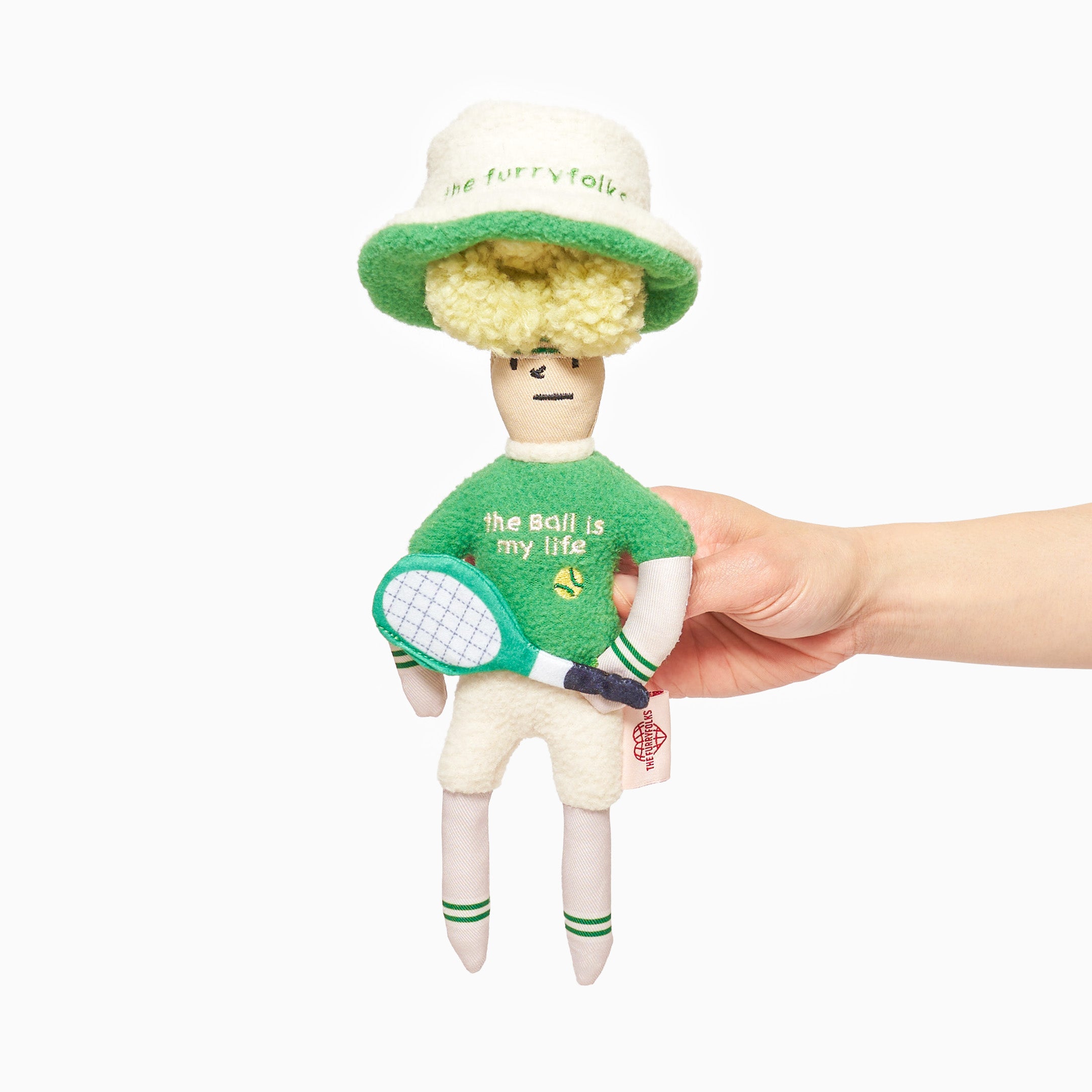  A hand is holding a plush toy styled as a tennis player, complete with a green hat labeled "the furryfolk," a green sweater with "the ball is my life" embroidered on it, and a white tennis racket. The toy has a simple, stitched face with a smile and eyes, and wears white, knee-high socks with green stripes. The design is whimsical and cartoonish.