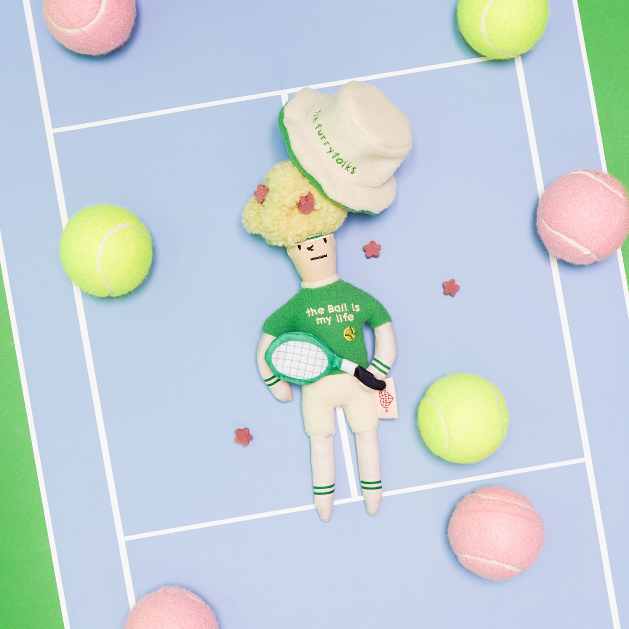 The image features a plush toy styled like a tennis player lying flat on a surface that resembles a tennis court, marked with blue and green lines. Surrounding the toy are various plush tennis balls in green and pink hues and small plush flower accents scattered about. The toy itself has a large, green hat labeled "the furryfolks," a green sweater with "the ball is my life" on the front, and holds a tennis racket. Its face is simple with stitched features, adding to the toy's playful charm.