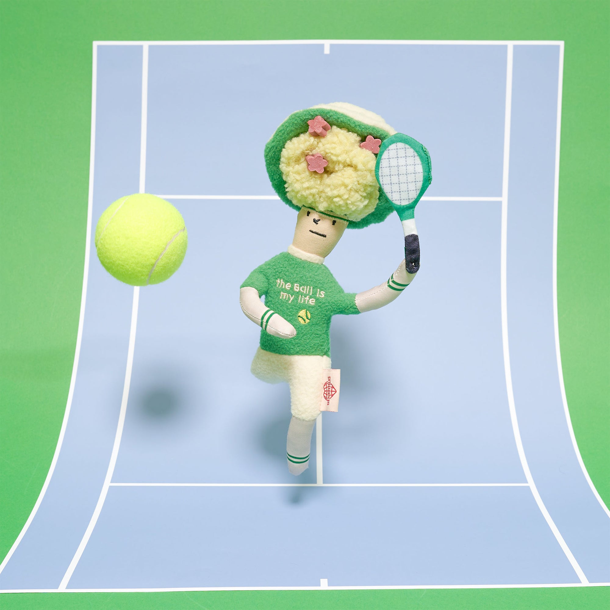 The toy is styled as a whimsical tennis player, complete with a green hat adorned with pink plush flowers, a green sweater with "the ball is my life" embroidered, and holding a white tennis racket. The figure has a stitched, simplistic face. A plush green tennis ball is depicted in motion, adding to the dynamic scene. The background is a graphic tennis court in vibrant greens, creating a playful and sporty setting.