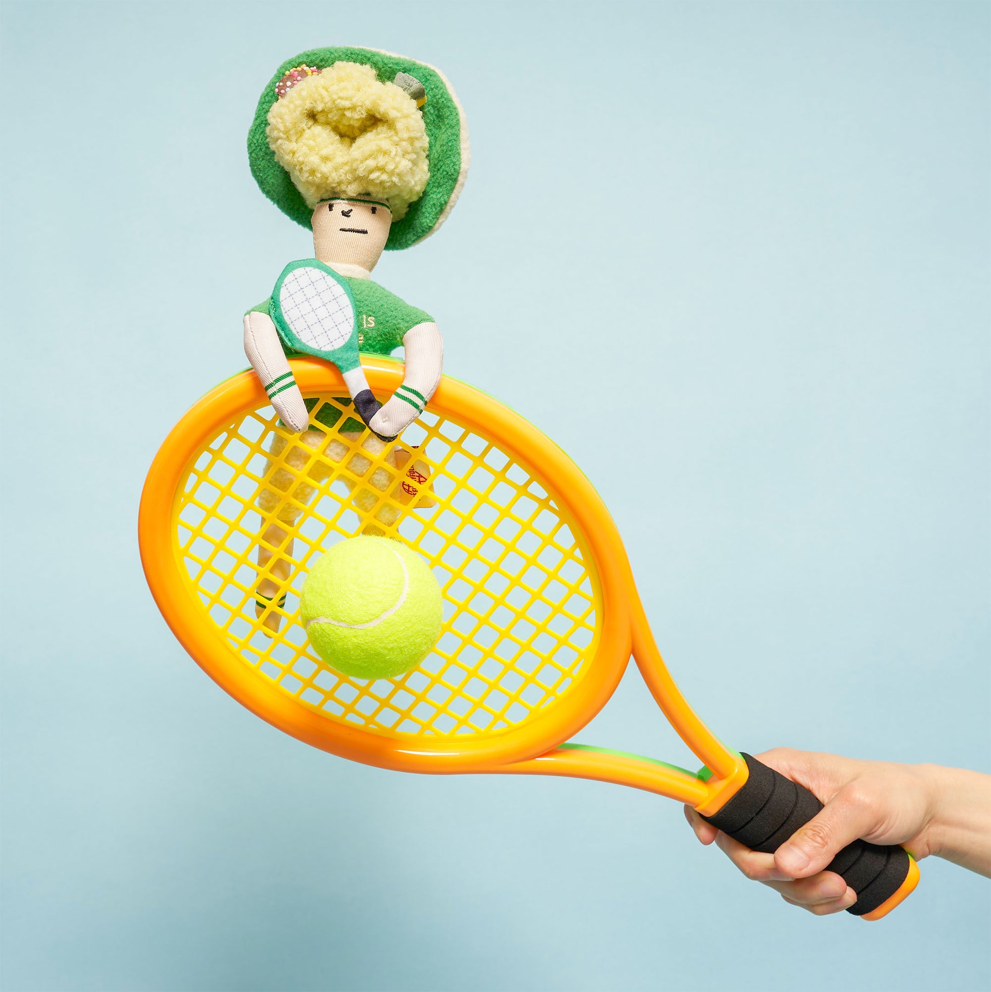 The image depicts a plush toy seated on an orange and yellow tennis racket held by a human hand. The toy is styled as a tennis player, with a green hat decorated with pink plush flowers, a green sweater with "the ball is my life" written on it, and a small white tennis racket. The background is a light blue, providing a clean and bright aesthetic.