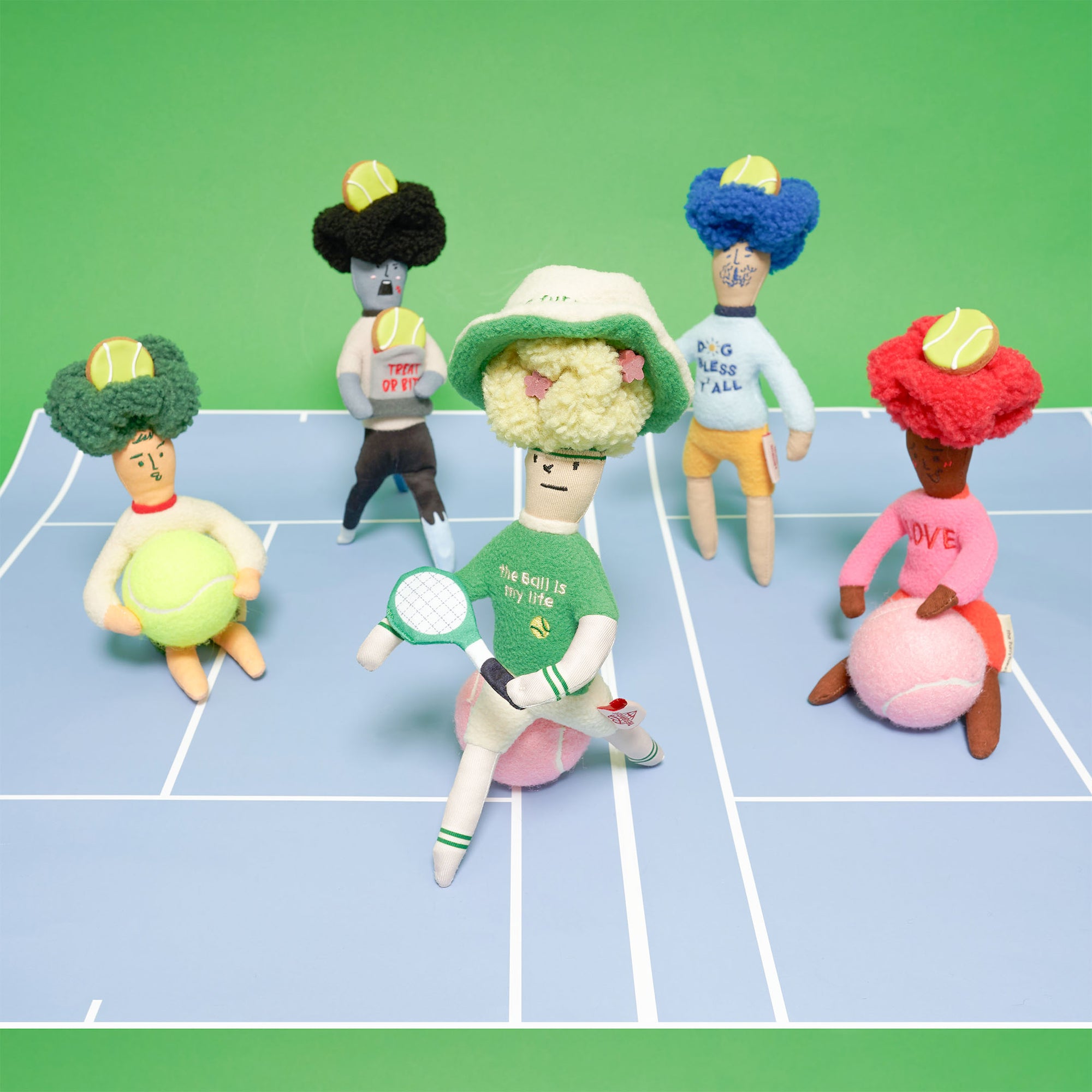 The image displays a collection of plush toys on a surface designed to look like a tennis court. Each toy is uniquely styled with different colors and outfits. The central toy wears a green sweater with "the ball is my life" and holds a tennis racket. Other toys include sweaters and hats in different colors. This scene is vibrant and playful, set against a backdrop of light green and blue, symbolizing the tennis court.