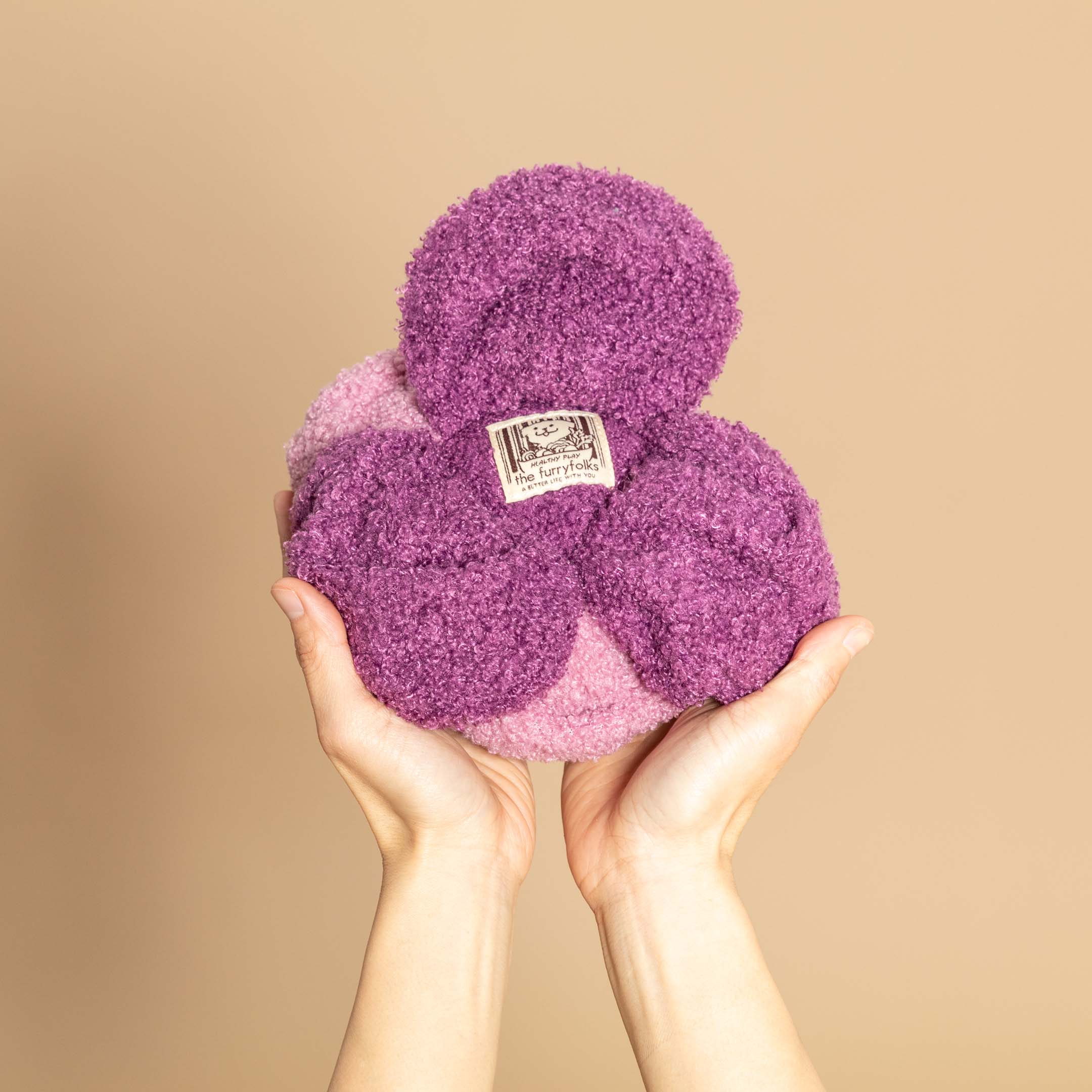 The image displays hands holding a purple and pink dog nosework toy resembling a red cabbage, with a "The Furryfolks" label. Designed for mental stimulation, the toy allows treats to be hidden within its layers, challenging a dog's sense of smell. The beige background contrasts with the vibrant toy.