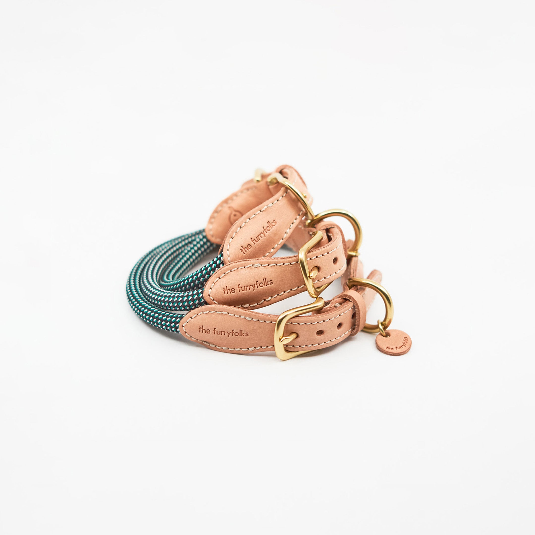 This image features a dog collar and leash set by "The Furryfolks". The pieces are fashioned with a teal and black woven fabric and complemented by tan leather accents and gold-tone hardware. Each leather piece is stamped with the brand's name, suggesting a signature design element. The background is white, which highlights the rich color and texture of the leash and collar. The design implies a blend of functionality and style for pet accessories.