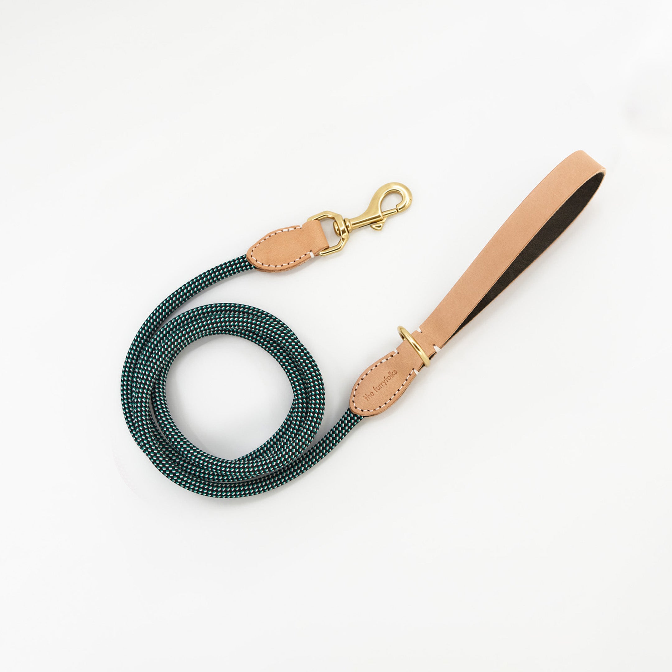 The image displays a teal and black woven dog leash with tan leather details and gold-tone metal hardware. It features a branded leather patch that reads "The Furryfolks," indicating its designer or brand. The leash is coiled, with the handle clearly visible, demonstrating its length and flexibility. The simple, clean white background ensures the focus is on the leash's elegant design and color scheme.