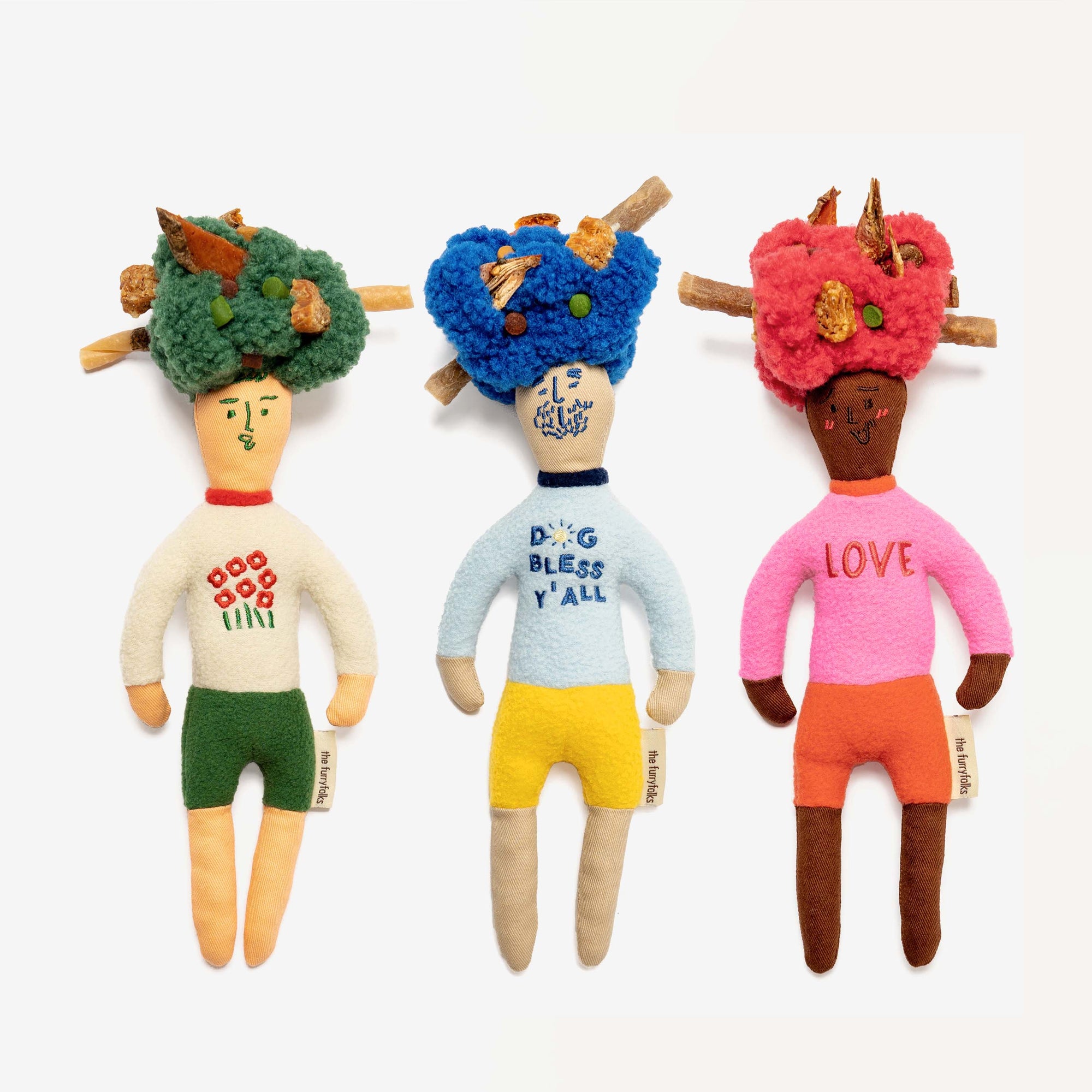 A collection of three human-shaped plush toys with colorful 'afro' hairstyles and shirts with phrases like 'Dog Bless Y'all' on a white surface.