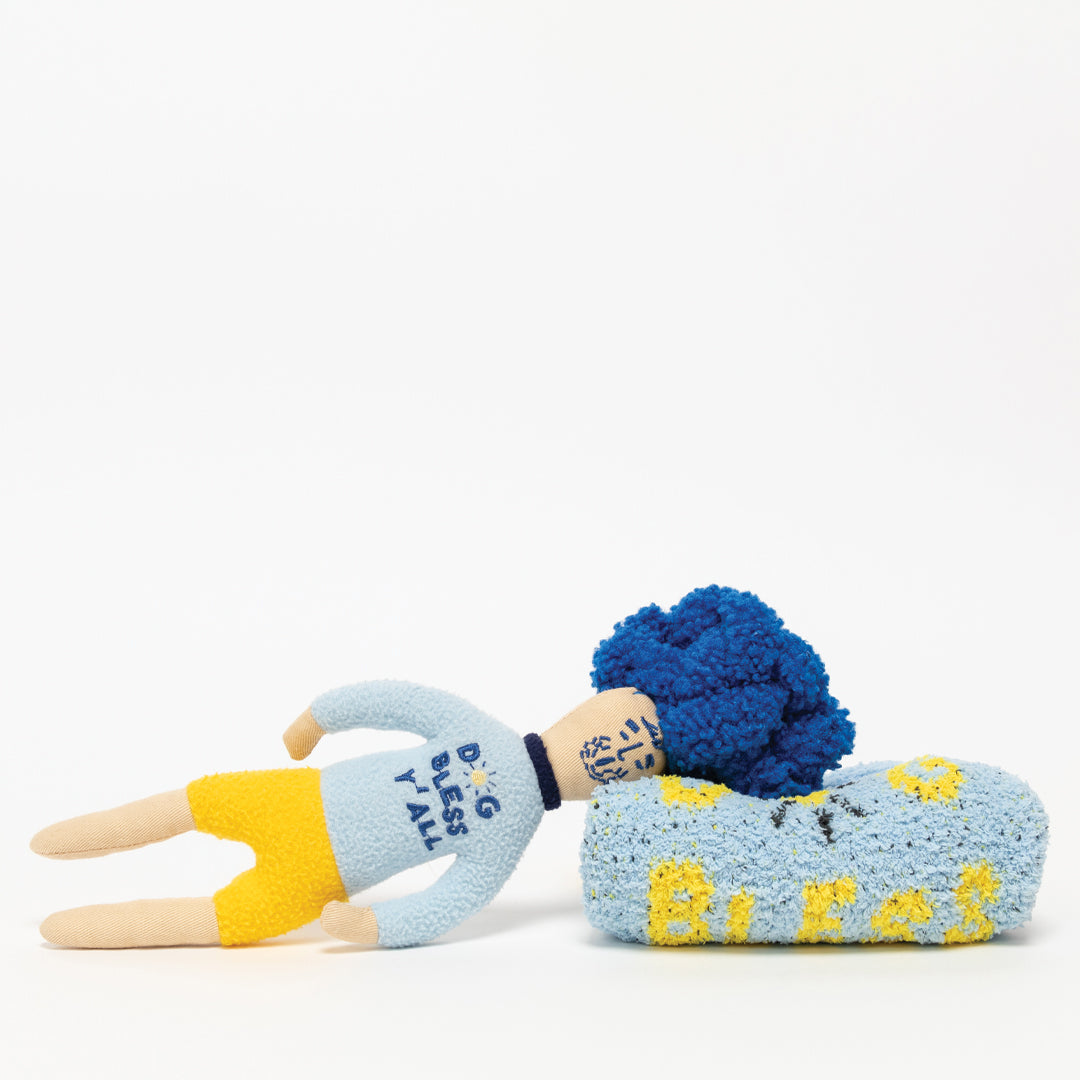 The image features two plush toys designed to resemble human items: one looks like a person with a blue body and yellow appendages wearing a shirt with the text "Dogs by Tali," and the other is a blue bed with a headrest and the word "DOG BLESS Y'ALL" written across. These whimsical toys are likely intended for pets and are crafted to mimic everyday objects, adding a humorous touch to the typical pet toy assortment.