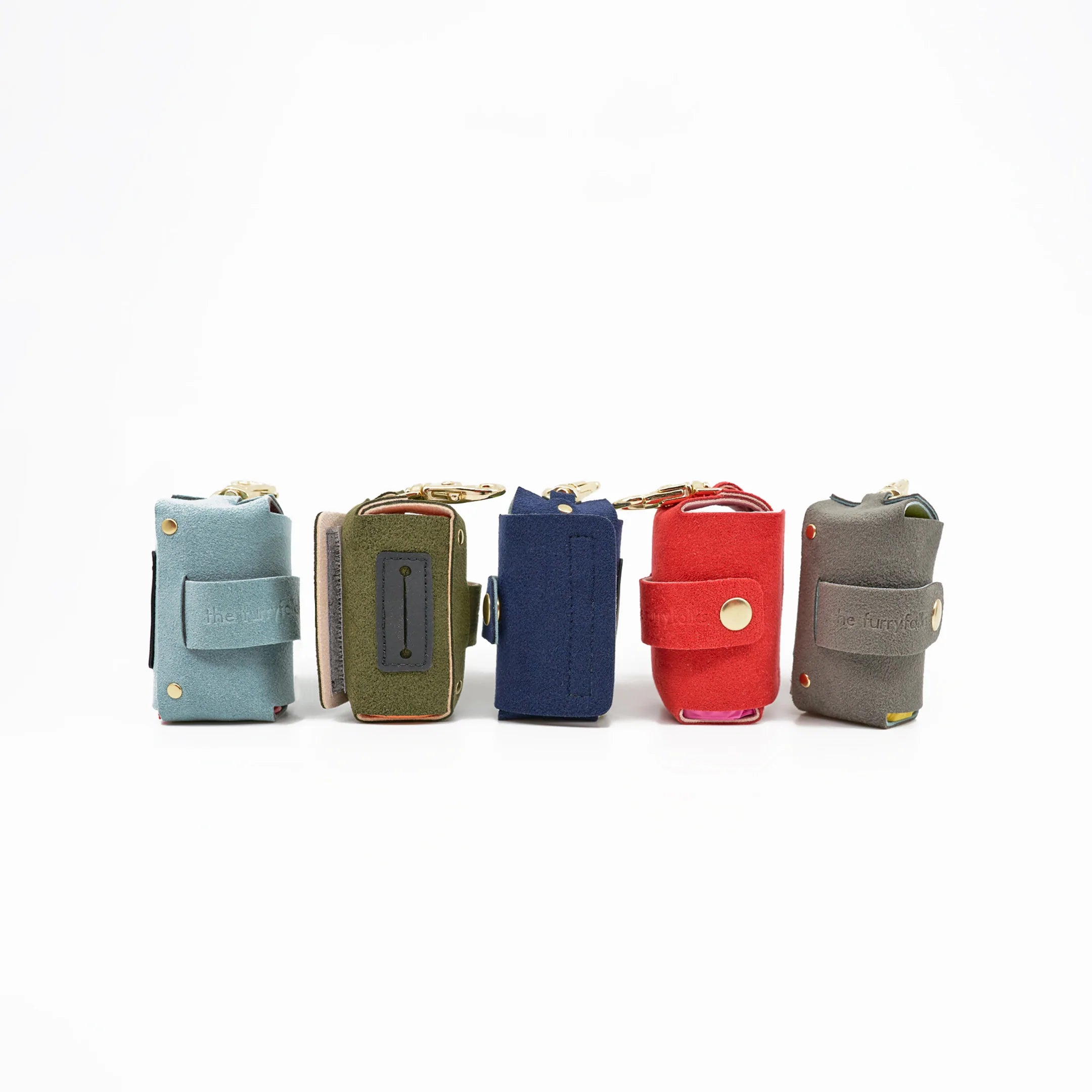 a lineup of five fabric dog poop bag holders in various colors. Each holder has a branded tag that reads "the furryfolks," likely indicating the manufacturer or brand of the product. These holders appear to be designed to attach to a dog's leash, providing a convenient and stylish way to carry poop bags while walking a dog. The variety of colors suggests that they are available to suit different tastes and preferences. 