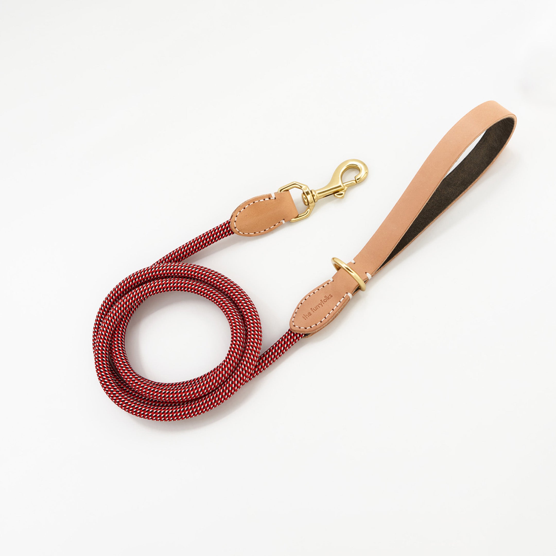 The image presents a red and white woven dog leash with tan leather accents and gold-tone hardware. A leather patch near the handle is embossed with "The Furryfolks," suggesting it is part of a pet accessory collection. The leash's design suggests durability and style, and the white background accentuates its bright colors and quality craftsmanship.